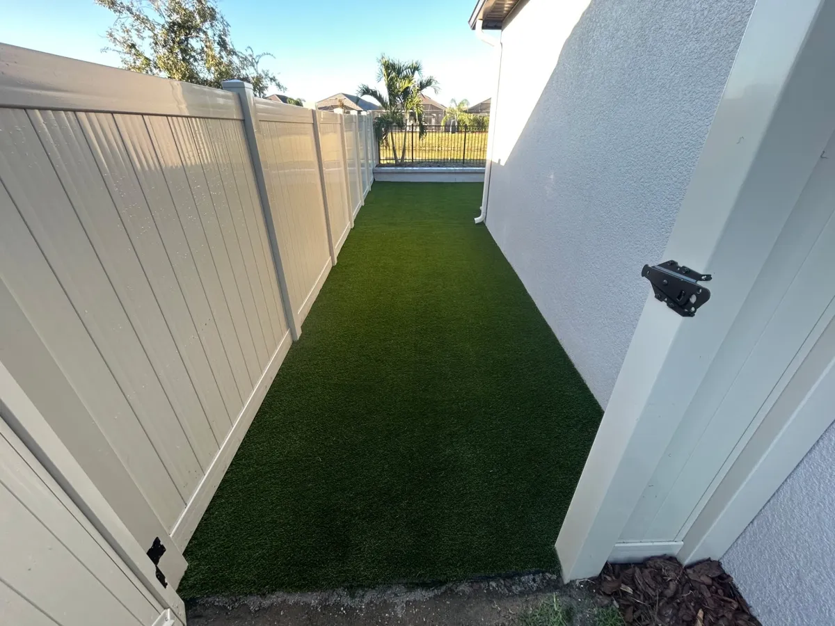 Narrow side yard covered in new artificial turf between a white fence and the house
